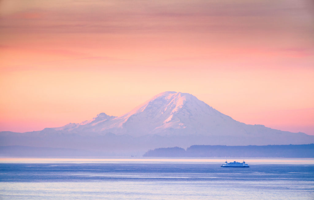 Looking over the Salish Sea/Puget Sound toward Tahoma/Mt. Rainier with a ferry in the distance, cool blue tones with a peach/orange sky, sunrise.