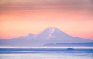 Looking over the Salish Sea/Puget Sound toward Tahoma/Mt. Rainier with a ferry in the distance, cool blue tones with a peach/orange sky, sunrise.