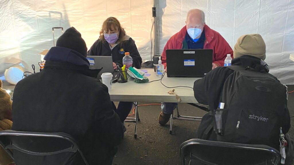 Two masked individuals on computers (both DOL staff), facing the camera, sitting across a table from two unidentified unhoused neighbors with their backs to the camera.