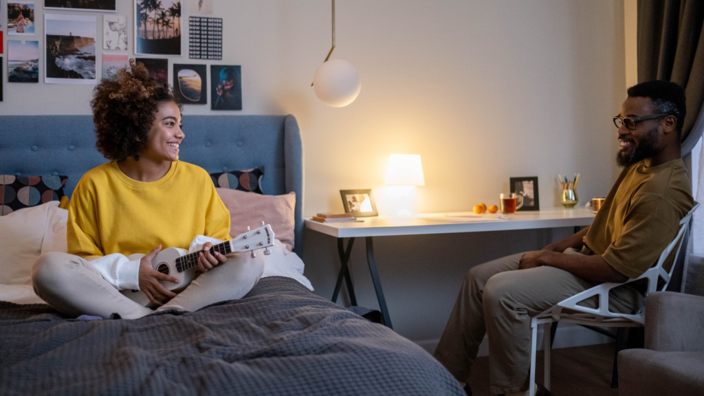 A woman sits on the bed playing the ukelele while a man sits at a desk next to her, they're both smiling in their safe, comfortable, small bedroom.