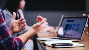 Hands in a position that indicates speaking with a laptop open with charts and graphs in front of the person speaking on the table.