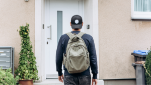A man with a backpack and a backwards hat approaches the door of an apartment building.