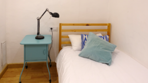 SRO-style shelter room with a twin bed, bedside table, and lamp.
